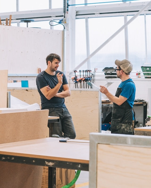 Two workers in a workshop discussing over wooden panels.